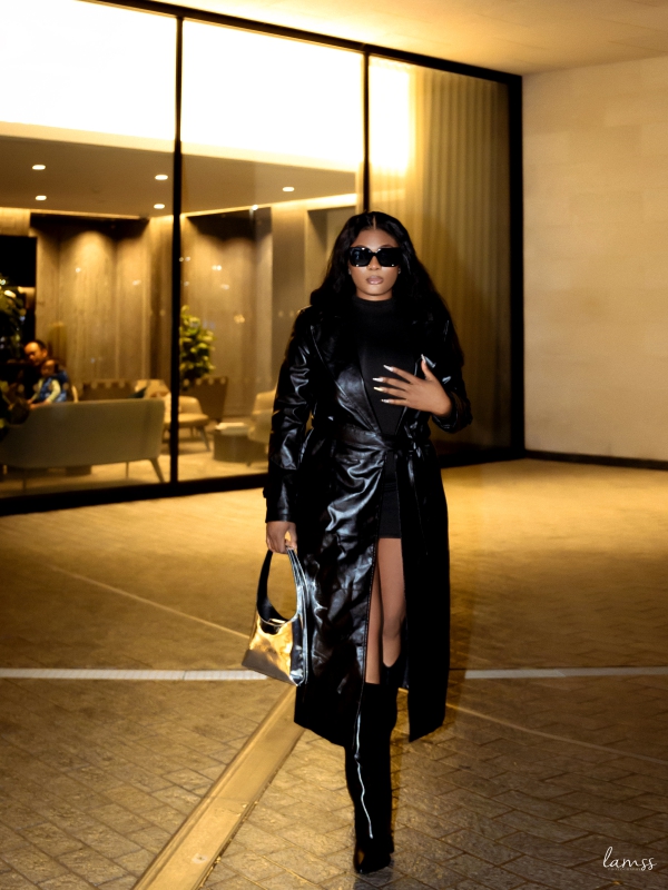 A Woman In A Long Black Leather Trench Coat And Sunglasses Walking Through A Modern Building Entrance.